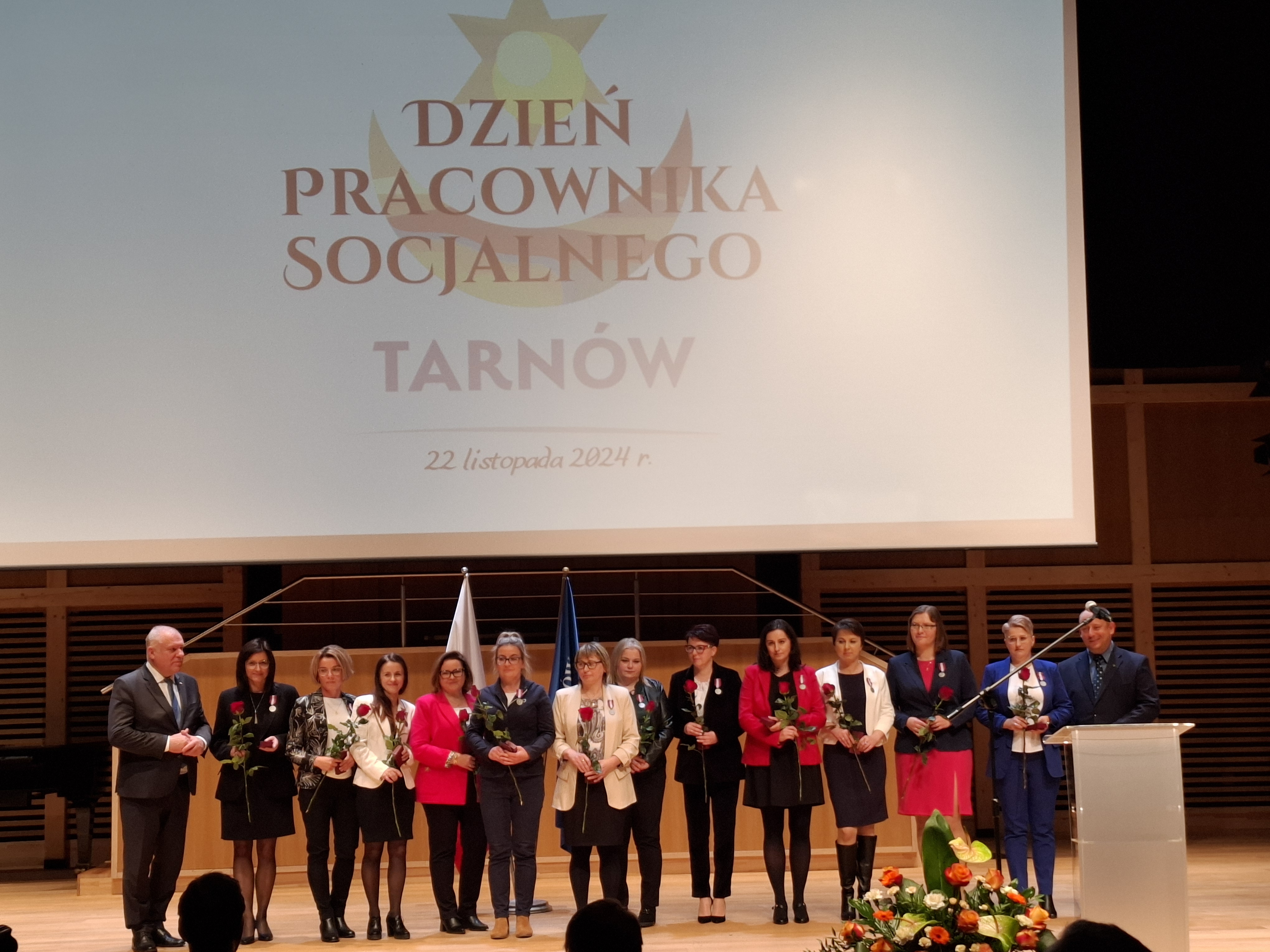 Grupowe zdjęcie pracowników socjalnych trzymających kwiaty, wszyscy stoją na scenie, nad nimi jest wyświetlany slajd z napisem Dzień Pracownika Socjalnego